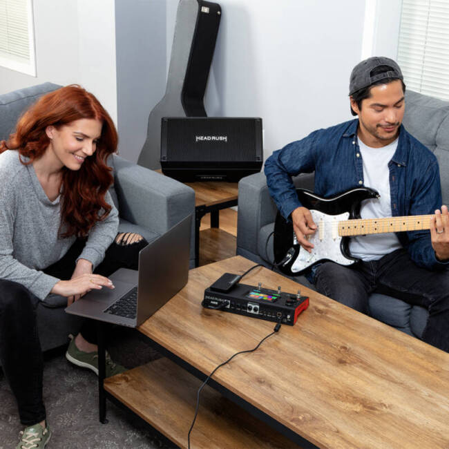 A man plays his electric guitar which is plugged into the HeadRush MX5 while a woman checks the output on her laptop