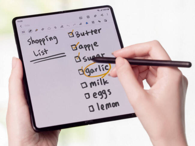 Samsung Galaxy Z Fold3 open with a shopping list showing; a woman's hand is writing with an S Pen Fold Edition stylus.