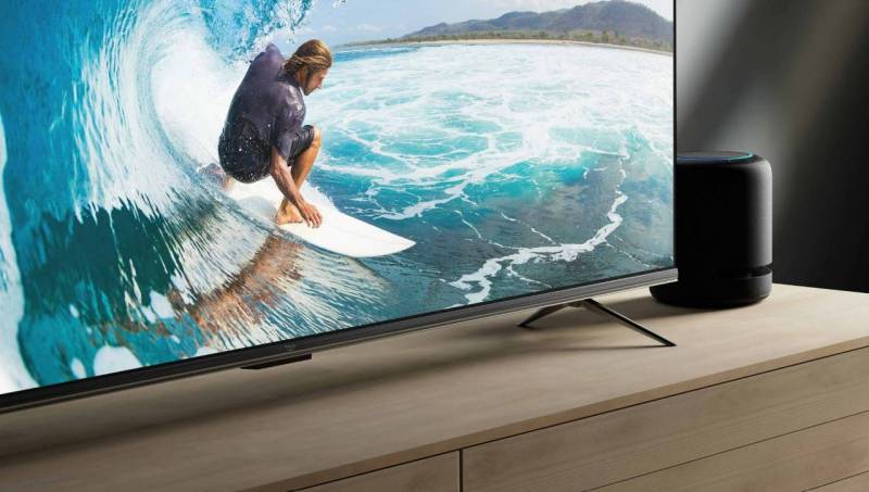 Amazon Fire TV Omni Series sits on a credenza next to a subwoofer. Onscreen, a man surfs in a wave.