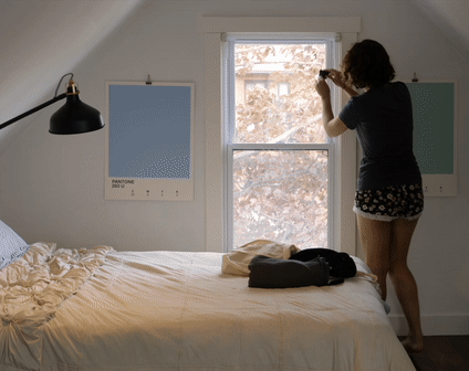 A woman hanging a Sleepout Curtain on her window with sunlight outside; the room is full dark once the curtain is hung