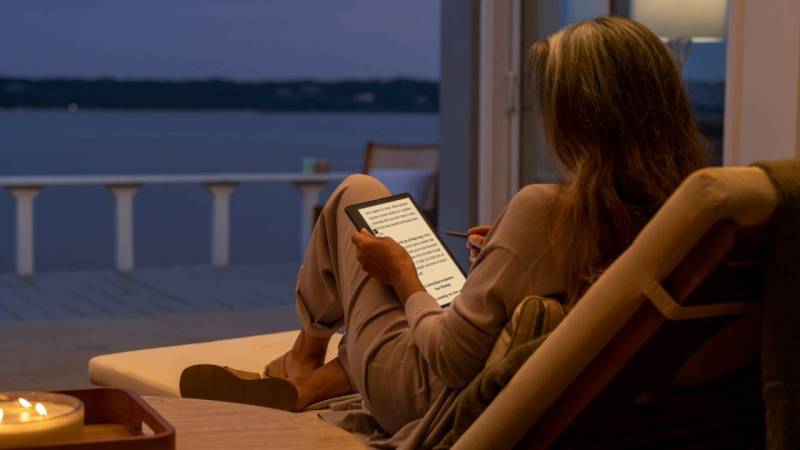 Woman making notes on her Amazon Kindle Scribe while relaxing by an open set of doors overlooking a lake.