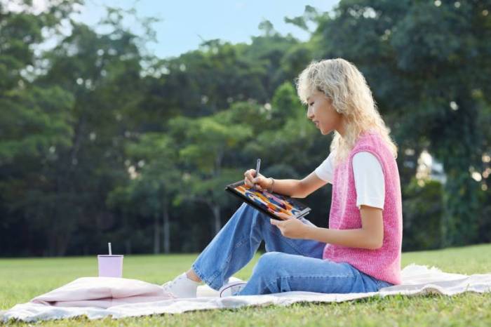 A woman wih blonde curlyhair wearing blue jeans, a white tee shirt, and a pink sweater vest uses a stylus to color on an LG Gram.