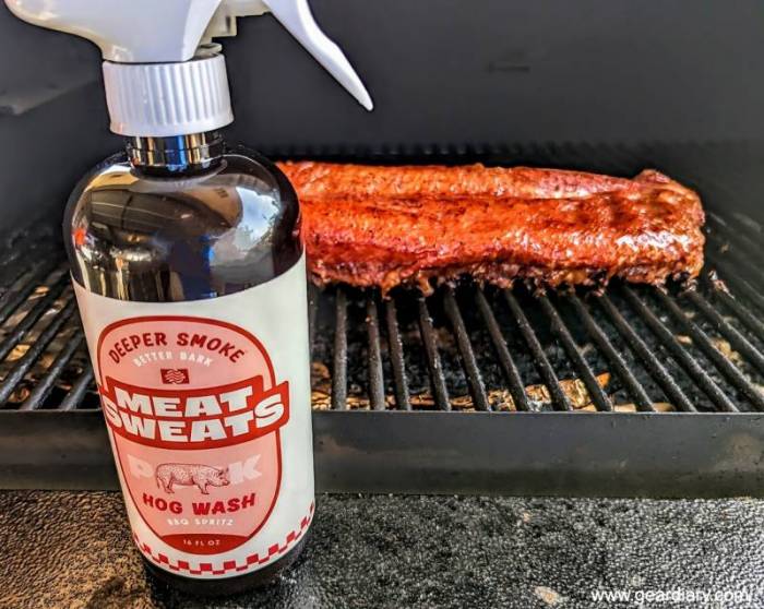 A spray bottle of Meat Sweats BBQ Spritz in Hog Wash flavor next to a smoking rack of pork ribs.