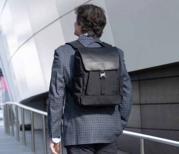 A man with dark hair walking around a building on a railed walkway looks forward while wearing a windowpane suit and a black on black WaterField Miles Laptop Backpack.