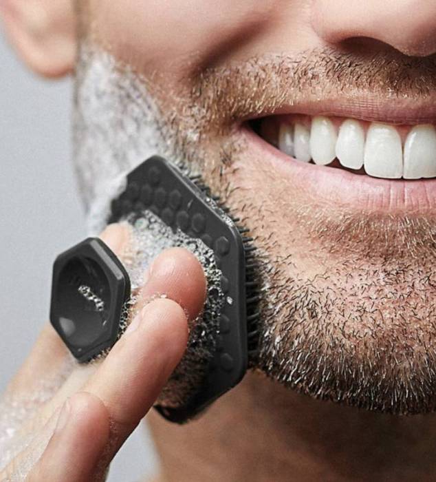 A person with a beard smiles as they use a Tooletries' face scrubber