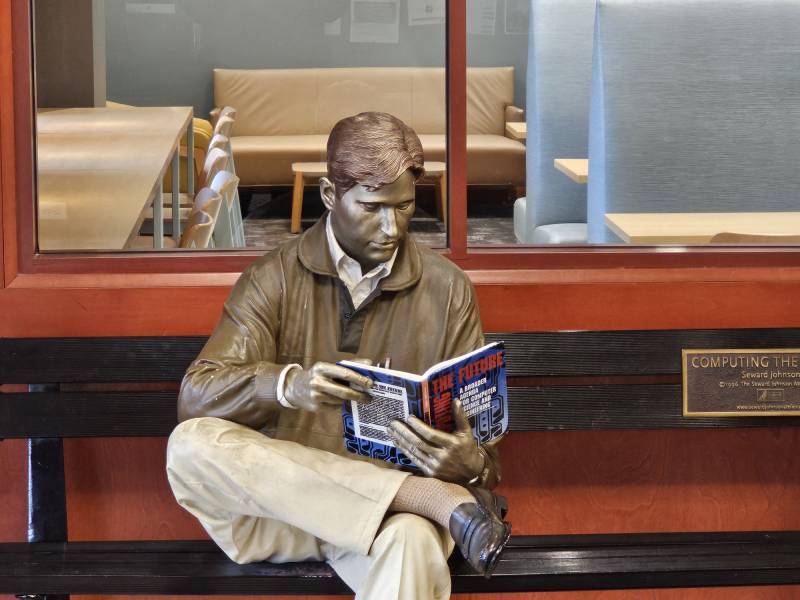 A statue of a man sits on a bench and reads a book.