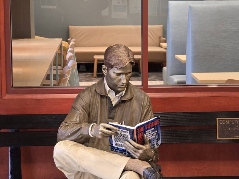 A statue of a man sits on a bench and reads a book.