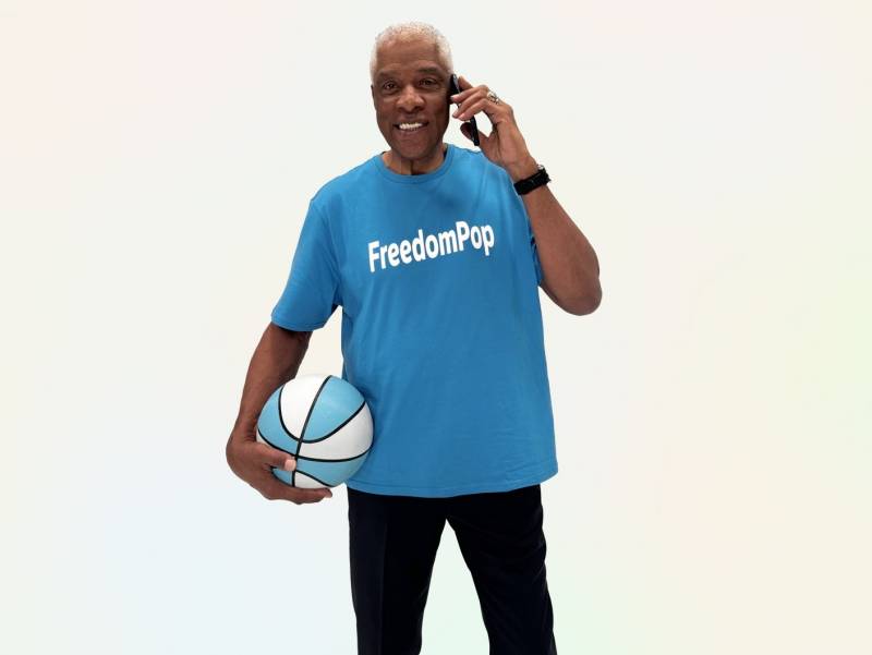 Dr J holding a basketball, wearing a blue FreedomPop t-shirt and holding a cellular phone to his ear
