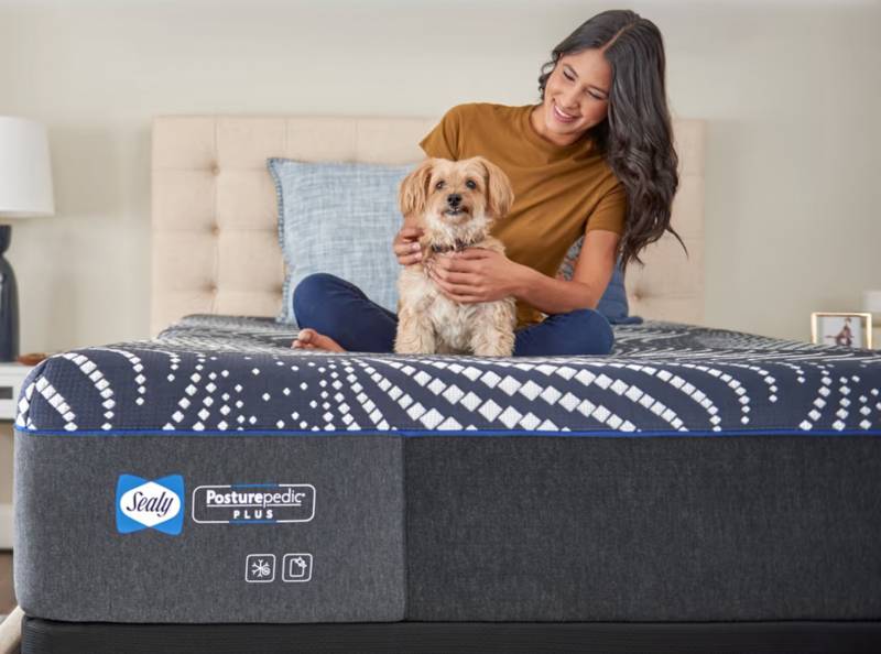 A woman and her dog sitting on the end of the Sealy Posturepedic Plus Hybrid mattress.
