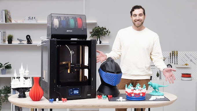 Man standing next to a Phrozen Arco 3D printer showing things he has printed, including a detailed castle, a vase, an armor helmet, a crown, and a T-Rex.