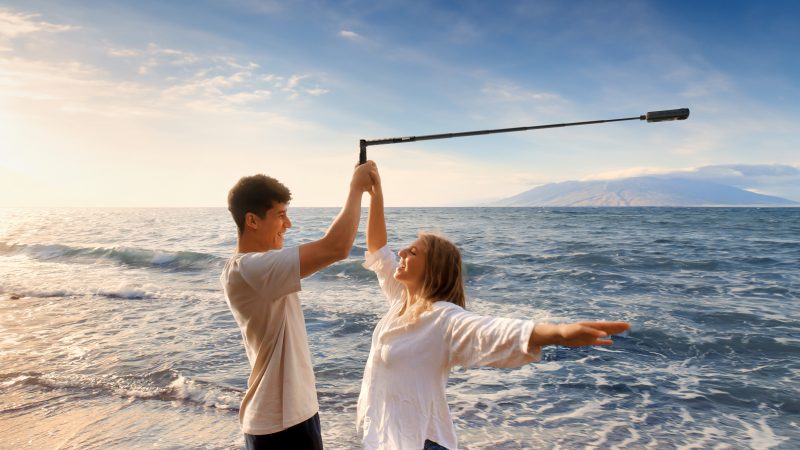 A couple stand in the water as the waves crash around them. The man is holding the Insta360 X4 on a selfie stick above the female with the selfie stick bent at an angle. 