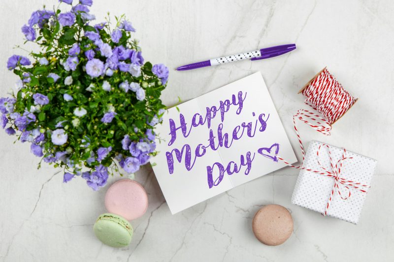 Happy Mother's Day photo of a card, purple flowers, macaroons, and a gift wrapped in white paper with a spool of red an white string next to it. 