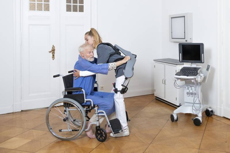 Woman assisting an elderly man from his wheelchair while wearing the Exia from German Bionics.