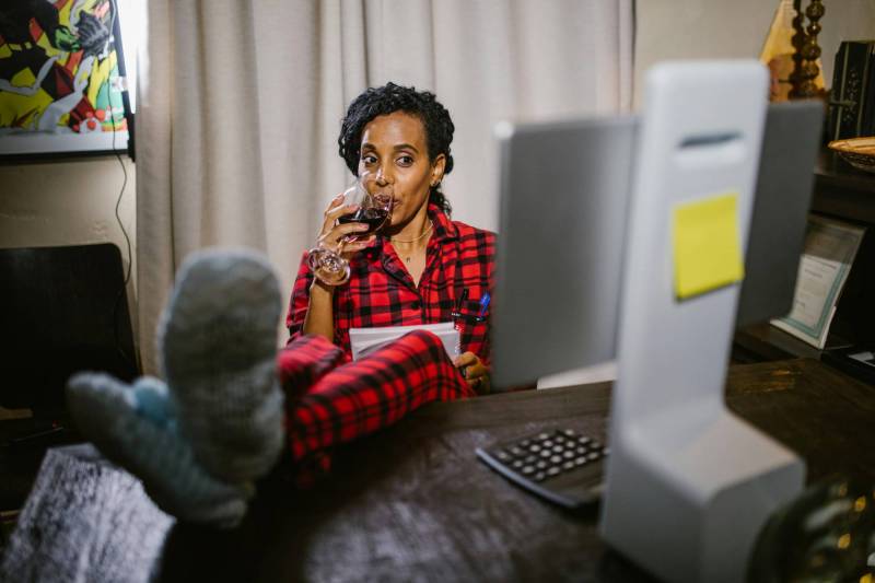 Photo of a woman, wearing pajamas, enjoying a glass of wine with her feet up at her desk.