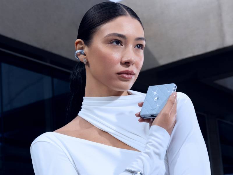 The Brilliant Collection - a woman with dark hair wearing a white dress holds the motorola razr in her right hand while wearing the moto buds loop in her right ear. 