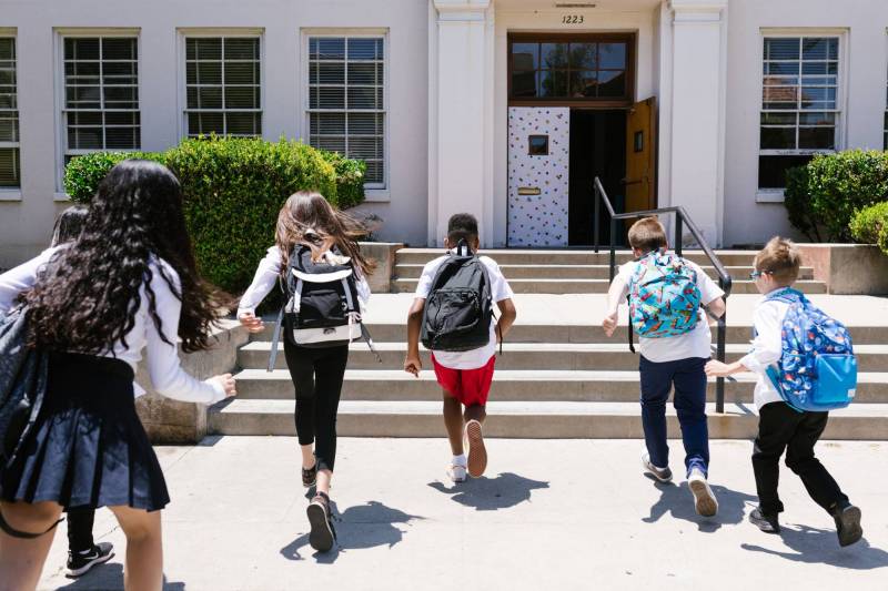 Gear Diary’s 2026 Back-to-School Gift Guide & Giveaway: Smart, Practical Picks for Every Stage of School and Beyond 1 Young children wearing backpacks run to the front of a school.